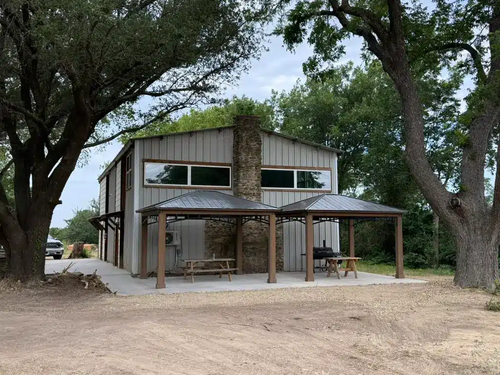 Metal barndominium building with stone chimney, two covered pavilions sheltering picnic tables and a barbecue smoker on