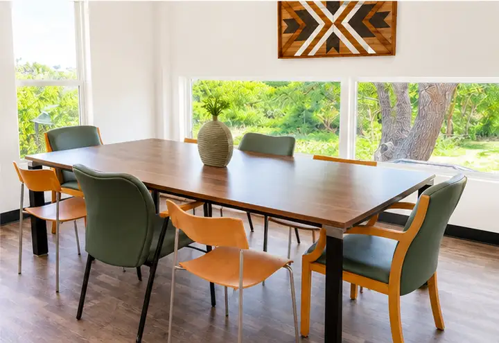 Indoor meeting room with long wooden conference table, green and orange upholstered chairs, vase centerpiece, and large
